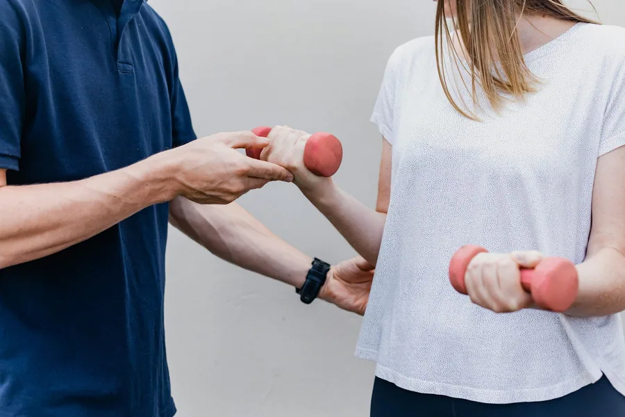 Ein Trainer führt den Arm einer Frau, die zwei kleine Hanteln hält.