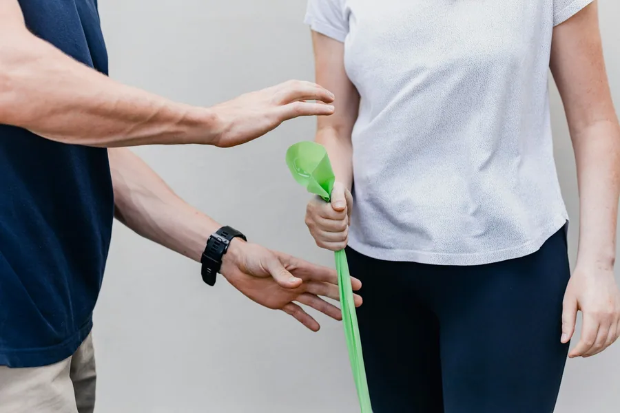 Eine Patientin hält ein grünes Therapieband, die Therapeutin korrigiert die Haltung ihres Arms.